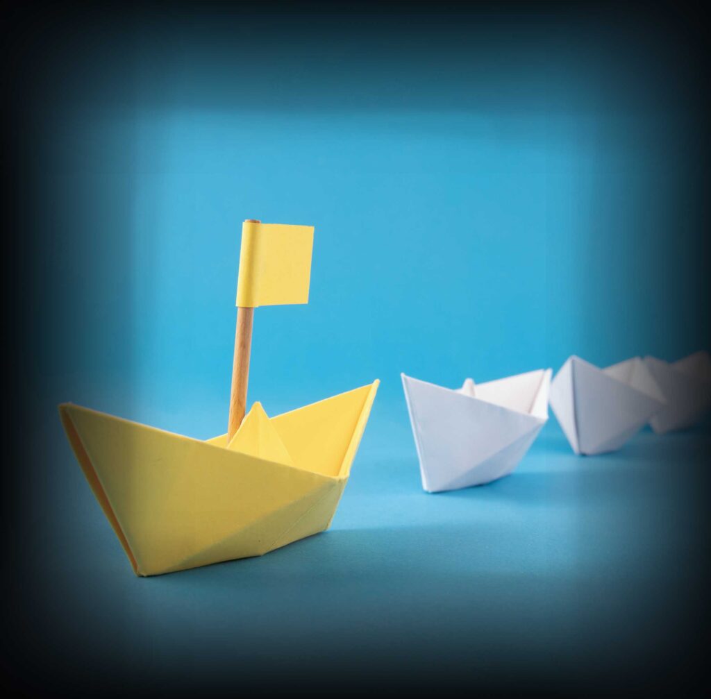Yellow Origami ship leading white origami ships on a blue background emulating leadership